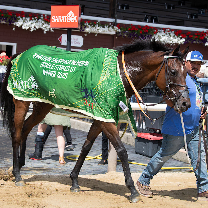 Horse wearing a green blanket with 'Saratoga' branding, being led by a person in a blue shirt and cap on a racetrack.